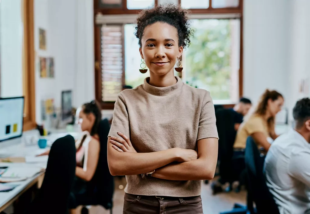Femme manager debout face à l’objectif, son équipe en train de travailler à l’arrière-plan.